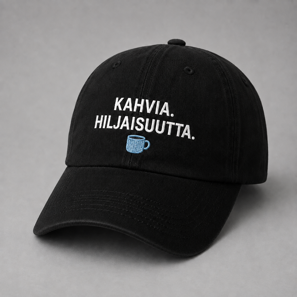 Musta kapealla kaarevalla lipalla varustettu lippis, jossa valkoinen Kahvia. Hiljaisuutta. -kirjonta ja vaaleansininen kahvikuppi