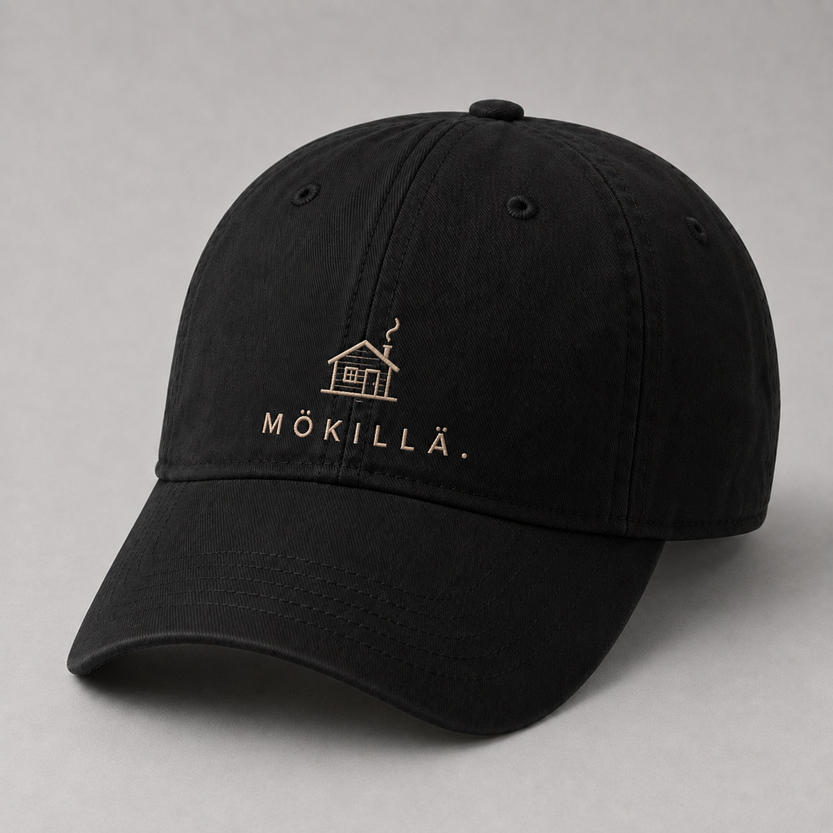Musta lippis, jossa vaalealla langalla brodeerattu pieni mökki ja teksti MÖKILLÄ.